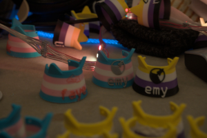 Verschiedene Katzenohren-Mate Tags in Trans und Nicht-Binär Design liegen verteilt auf einem Tisch, mit anderem Krimskrams und einer Lichterkette darauf.