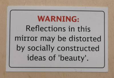 Weißer Sticker mit rotem Text "WARNING:", darunter in Schwarz "Reflections in this mirror may be distorted by socially constructed ideas of 'beauty'.", drum herum ein schwarzer Rahmen mit abgerundeten Ecken. Der Sticker liegt auf einem Holzbrett. Weißer Sticker mit rotem Text "WARNING:", darunter in Schwarz "Reflections in this mirror may be distorted by socially constructed ideas of 'beauty'.", drum herum ein schwarzer Rahmen mit abgerundeten Ecken. Der Sticker liegt auf einem Holzbrett.